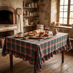 Könnte beinhalten: Ein rechteckiger Tisch, der mit einer rot-grün-weiß karierten Tischdecke mit Rüschenrand bedeckt ist. Auf dem Tisch stehen ein hölzernes Schneidebrett mit Brot und Croissants, eine Keramiktasse, ein Glas Marmelade und ein kleiner Keramikkrug.