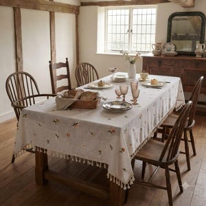 May include: A rectangular off-white tablecloth featuring small embroidered floral motifs in orange and green, finished with a decorative fringe along the edges. The cloth is draped over a wooden dining table surrounded by four wooden spindle chairs.
