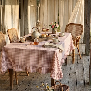 May include: A rectangular table covered with a pink and white gingham tablecloth featuring a ruffled edge. The table is set with a ceramic teapot, a wooden tray holding a tiered dessert stand with pastries, teacups, and a honey jar with a dipper.