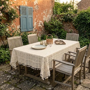 May include: A rectangular off-white crochet tablecloth with a repeating diamond pattern and fringe trim, draped over a wooden outdoor dining table surrounded by four wooden slatted chairs, featuring a woven basket, tray, and glasses on top.