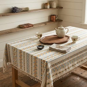 May include: A rectangular tablecloth featuring a cream base with vertical stripes of geometric patterns in muted blue, mustard yellow, and brown. The edges are finished with short, cream-colored fringe tassels along the entire perimeter.