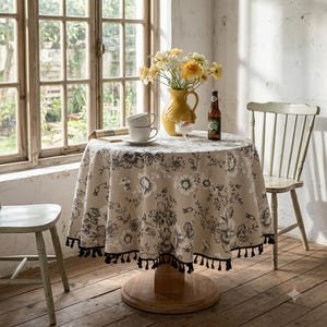 Könnte beinhalten: Ein runder Tisch mit einer beigen Tischdecke mit schwarzem Blumenmuster und schwarzer Quastenborte. Auf dem Tisch stehen ein gelber Keramikkrug mit gelben und weißen Blumen, zwei weiße Keramiktassen und eine braune Glasflasche.