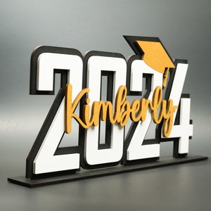 May include: White and black wooden 2024 graduation sign with a gold graduation cap and the name Kimberly.