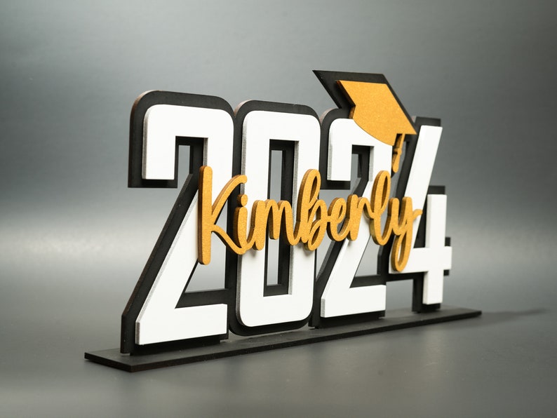 May include: White and black wooden 2024 graduation sign with a gold graduation cap and the name Kimberly.