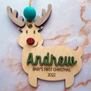 May include: Wooden reindeer Christmas ornament with a green and white name "Andrew" and the text "Baby's First Christmas 2022". The reindeer has a red glitter nose and a turquoise bead at the top.