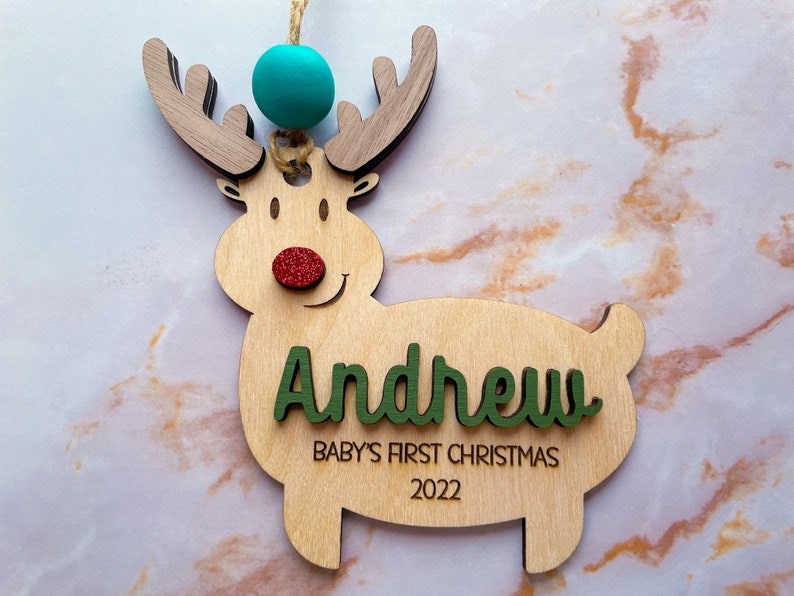 May include: Wooden reindeer Christmas ornament with a green and white name "Andrew" and the text "Baby's First Christmas 2022". The reindeer has a red glitter nose and a turquoise bead at the top.