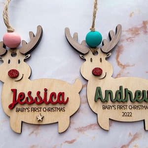 May include: Two wooden reindeer ornaments with glitter noses and red and green names. The reindeer on the left has a pink bead and the text "Jessica Baby's First Christmas" and the reindeer on the right has a turquoise bead and the text "Andrew Baby's First Christmas 2022".