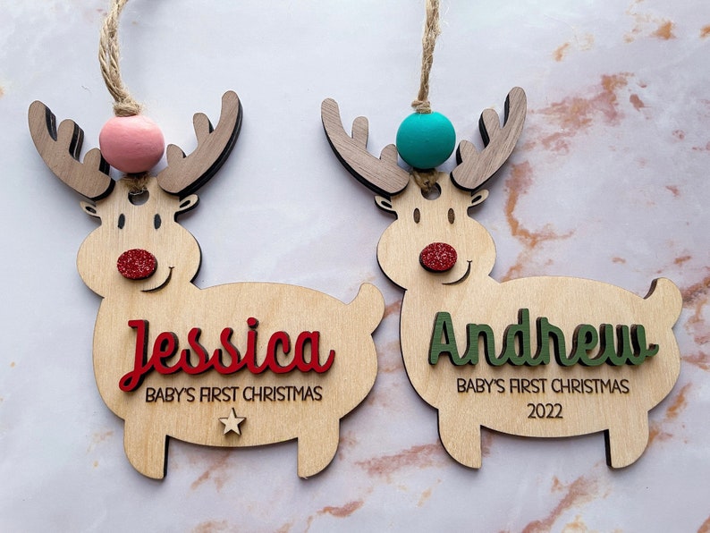 May include: Two wooden reindeer ornaments with glitter noses and red and green names. The reindeer on the left has a pink bead and the text "Jessica Baby's First Christmas" and the reindeer on the right has a turquoise bead and the text "Andrew Baby's First Christmas 2022".