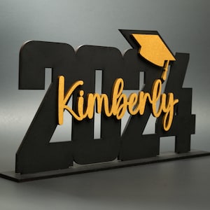 May include: Black wooden 2024 graduation sign with a gold graduation cap and the name Kimberly in gold lettering.