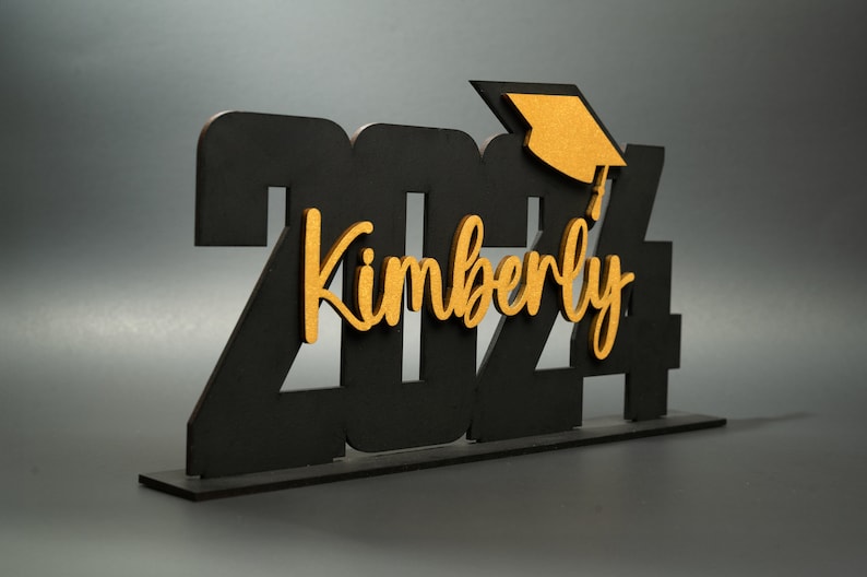 May include: Black wooden 2024 graduation sign with a gold graduation cap and the name Kimberly in gold lettering.