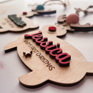 May include: A wooden reindeer ornament with a red "Jessica" name and "Baby's First Christmas" text. The ornament has a white star and a red pom-pom.
