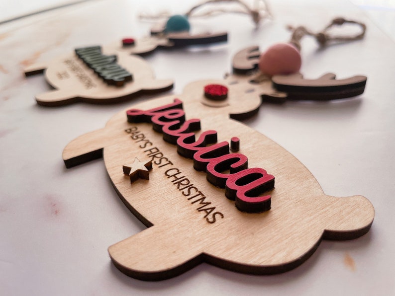 May include: A wooden reindeer ornament with a red "Jessica" name and "Baby's First Christmas" text. The ornament has a white star and a red pom-pom.