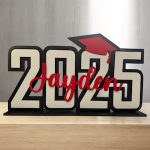 May include: A black and white wooden sign with the year 2025 and the name Jayden in red lettering. A red graduation cap is on top of the sign.