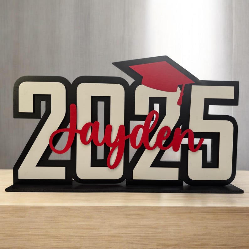 May include: A black and white wooden sign with the year 2025 and the name Jayden in red lettering. A red graduation cap is on top of the sign.
