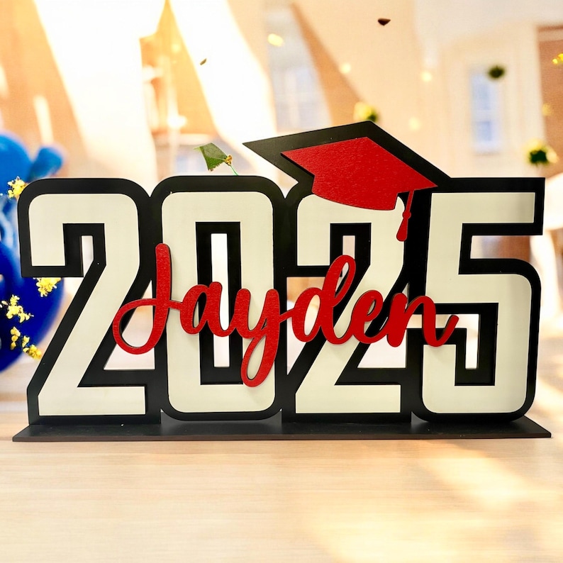 May include: Black and white wooden sign with the year 2025 and the name Jayden in red lettering. A red graduation cap is on top of the sign.