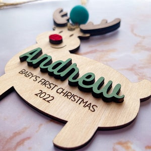 May include: Wooden reindeer ornament with a green "Andrew" name and "Baby's First Christmas 2022" text. The reindeer has a red nose and a blue and green pom-pom on its antlers.