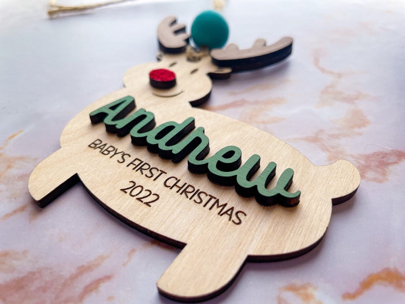 May include: Wooden reindeer ornament with a green "Andrew" name and "Baby's First Christmas 2022" text. The reindeer has a red nose and a blue and green pom-pom on its antlers.