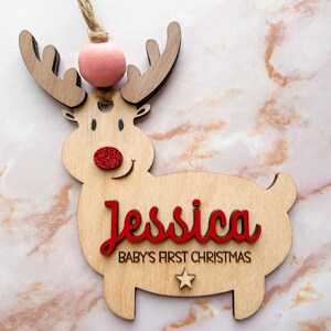 May include: Wooden reindeer ornament with a pink pom-pom on top, red glitter nose, and the name "Jessica" and the text "Baby's First Christmas" in red lettering.