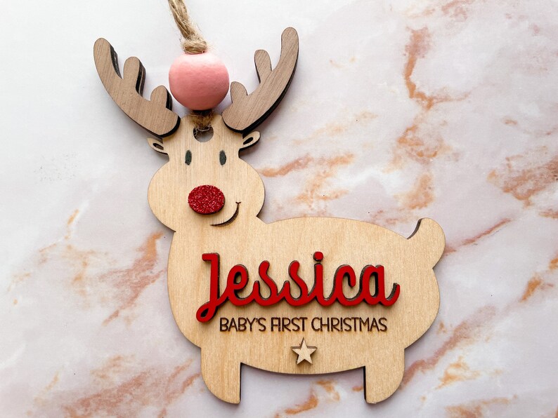 May include: Wooden reindeer ornament with a pink pom-pom on top, red glitter nose, and the name "Jessica" and the text "Baby's First Christmas" in red lettering.