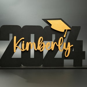 May include: Black wooden 2024 graduation sign with a gold graduation cap and the name Kimberly in gold lettering.