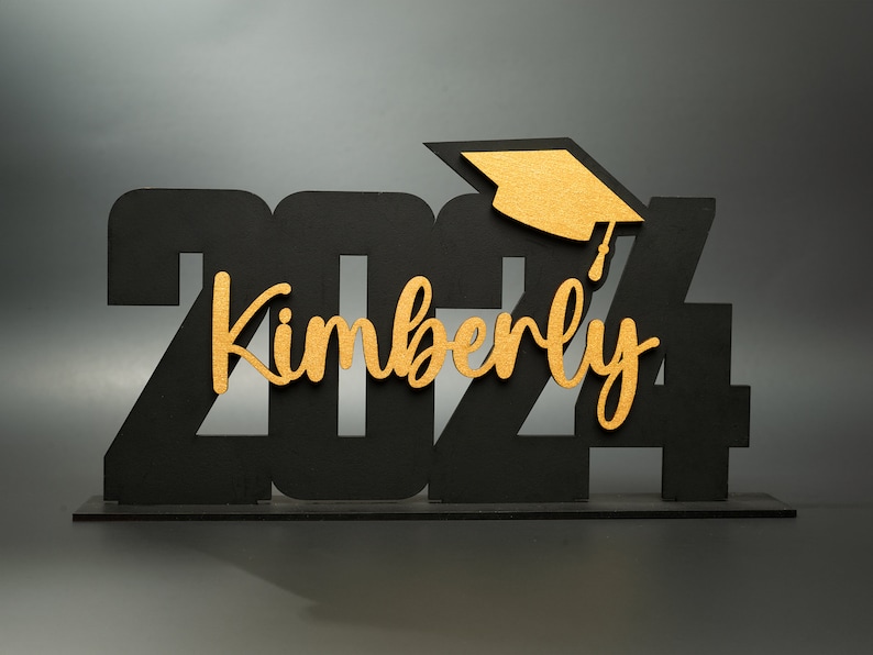 May include: Black wooden 2024 graduation sign with a gold graduation cap and the name Kimberly in gold lettering.