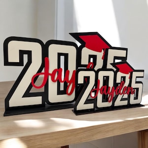 May include: Black and white wooden cutout of the year 2025 with a red graduation cap and the name "Jayden" in red cursive lettering.