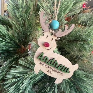 May include: A wooden reindeer Christmas ornament with a green name "Andrew" and the text "Baby's First Christmas 2022". The reindeer has a red glitter nose and a blue bead at the top.