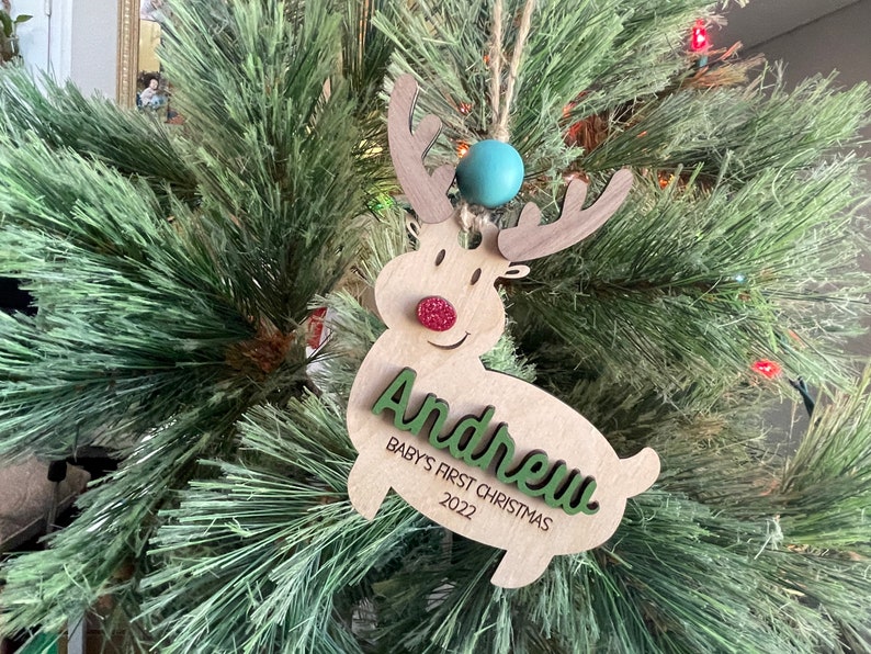 May include: A wooden reindeer Christmas ornament with a green name "Andrew" and the text "Baby's First Christmas 2022". The reindeer has a red glitter nose and a blue bead at the top.