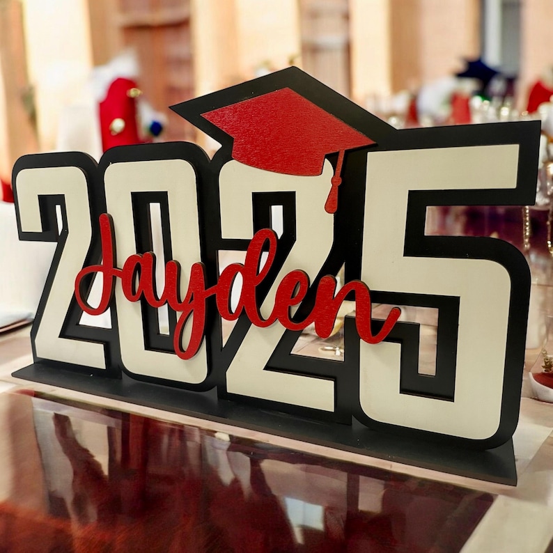 May include: A white and black wooden tabletop sign with the year 2025 and the name Jayden in red lettering. A red graduation cap is on top of the sign.