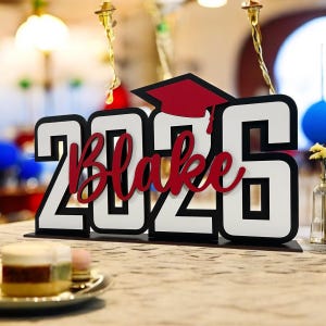 Könnte beinhalten: Ein Holzschild zum Thema Abschluss mit dem Namen "Blake" in roter Schrift über dem Jahr "2026". Die Zahlen sind weiß mit schwarzen Umrissen, und eine rote Abschlusskappe befindet sich über der "26". Das Schild steht auf einem Tisch, im Vordergrund ist ein Dessert zu sehen.
