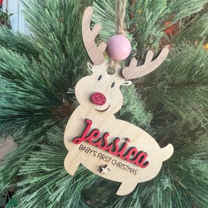 May include: A wooden reindeer Christmas ornament with a red nose and glitter accents. The ornament is personalized with the name "Jessica" and the text "Baby's First Christmas".