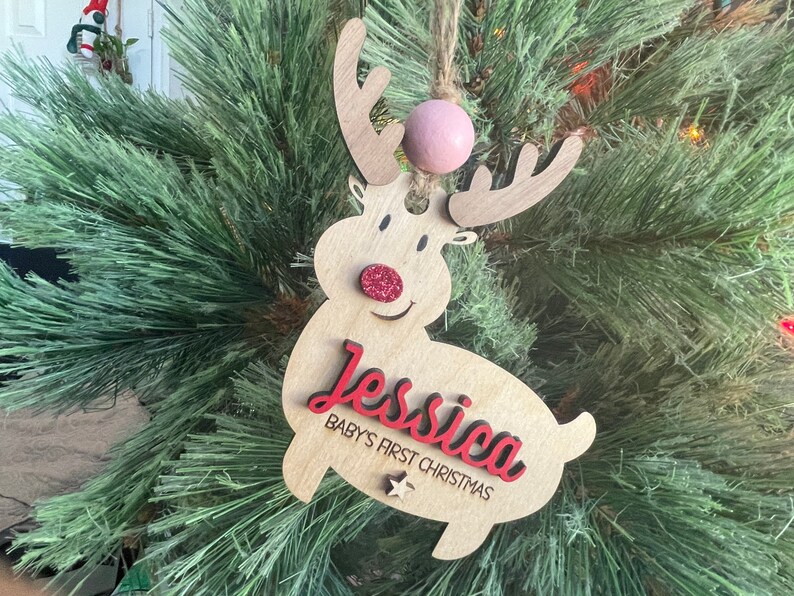 May include: A wooden reindeer Christmas ornament with a red nose and glitter accents. The ornament is personalized with the name "Jessica" and the text "Baby's First Christmas".