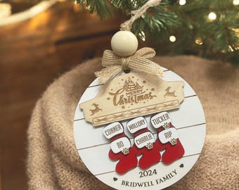 May include: A round wooden Christmas ornament with a white background and a red and white stocking for each family member. The ornament features the names Conner, Bo, Mallory, Charlie, Tucker, Rip, and the text "Merry Christmas", "2024", and "Bridwell Family".