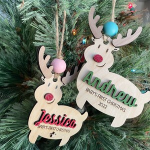 May include: Two wooden reindeer ornaments with the names "Jessica" and "Andrew" on them. The ornaments are decorated with glitter and have a red nose and a pink or blue bead on the top. The text on the ornaments reads "Baby's First Christmas 2022".