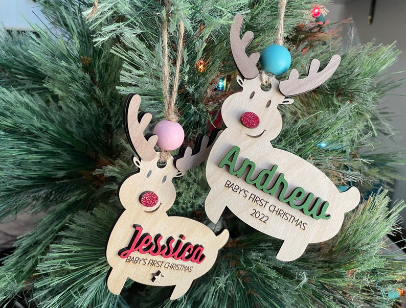 May include: Two wooden reindeer ornaments with the names "Jessica" and "Andrew" on them. The ornaments are decorated with glitter and have a red nose and a pink or blue bead on the top. The text on the ornaments reads "Baby's First Christmas 2022".