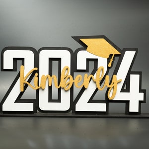 May include: A black and white 2024 graduation sign with a gold graduation cap and the name Kimberly in gold lettering.