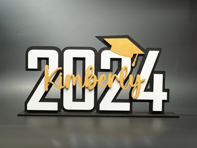 May include: A black and white 2024 graduation sign with a gold graduation cap and the name Kimberly in gold lettering.