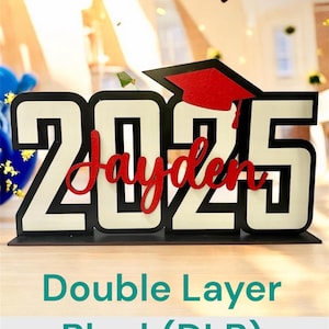May include: A black and white layered wooden sign with the year "2025" and the name "Jayden" in red. The sign has a graduation cap on top and is on a wooden surface. The text "Double Layer Black (DLB)" is below the sign.