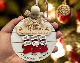 May include: A white wooden Christmas ornament with a red and white stocking design. The ornament features the names Jon, Megan, Alina, Audrey, Riley, and Piper, along with the text "Merry Christmas", "2024", and "The Sfrengeue Family".