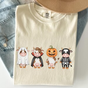 May include: Cream-colored Comfort Colors t-shirt featuring four cartoon cows dressed in Halloween costumes: a ghost, a vampire, a jack-o'-lantern, and a skeleton. The shirt is paired with blue jeans and a necklace.