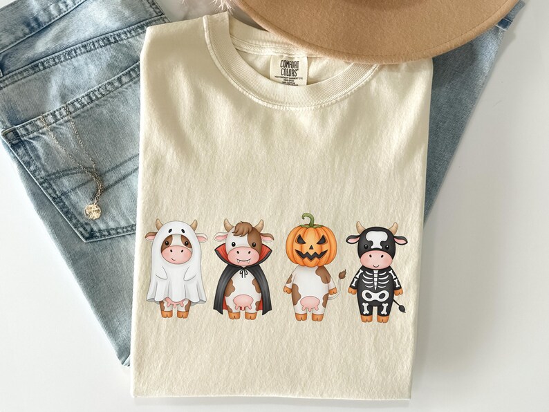May include: Cream-colored Comfort Colors t-shirt featuring four cartoon cows dressed in Halloween costumes: a ghost, a vampire, a jack-o'-lantern, and a skeleton. The shirt is paired with blue jeans and a necklace.