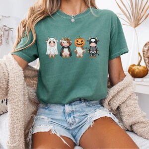 May include: A teal green t-shirt featuring four cartoon cows dressed in Halloween costumes: a ghost, a vampire, a pumpkin, and a skeleton. The shirt is worn with denim shorts and a cream-colored cardigan.