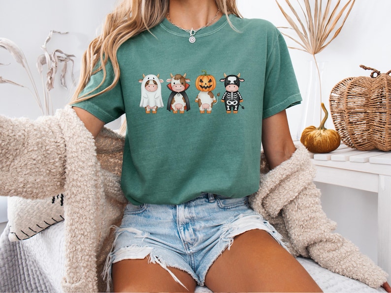 May include: A teal green t-shirt featuring four cartoon cows dressed in Halloween costumes: a ghost, a vampire, a pumpkin, and a skeleton. The shirt is worn with denim shorts and a cream-colored cardigan.