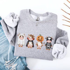 May include: A heather gray sweatshirt featuring four cartoon cows dressed in Halloween costumes: a ghost, a vampire, a jack-o'-lantern, and a skeleton. The sweatshirt is paired with blue jeans on a white surface.