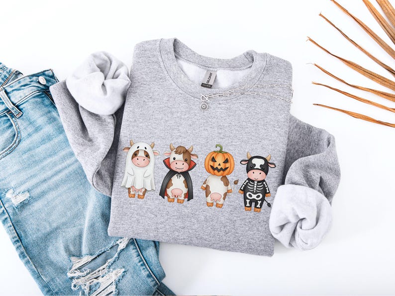 May include: A heather gray sweatshirt featuring four cartoon cows dressed in Halloween costumes: a ghost, a vampire, a jack-o'-lantern, and a skeleton. The sweatshirt is paired with blue jeans on a white surface.