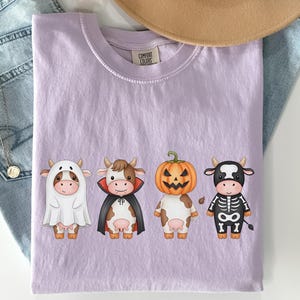 May include: A light purple Comfort Colors t-shirt featuring four cartoon cows dressed in Halloween costumes: a ghost, a vampire, a jack-o'-lantern, and a skeleton. The shirt is displayed on a white surface with a pair of blue jeans and a tan hat.