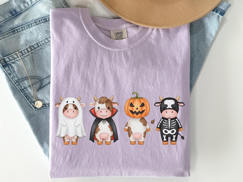 May include: A light purple Comfort Colors t-shirt featuring four cartoon cows dressed in Halloween costumes: a ghost, a vampire, a jack-o'-lantern, and a skeleton. The shirt is displayed on a white surface with a pair of blue jeans and a tan hat.