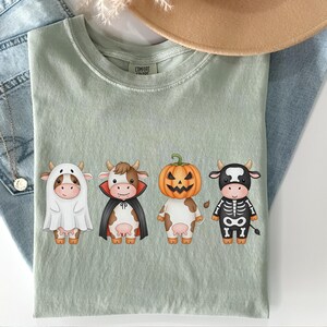 May include: Sage green t-shirt featuring four cartoon cows dressed in Halloween costumes: a ghost, a vampire, a jack-o'-lantern, and a skeleton. The shirt is laid on top of blue jeans and a tan hat.