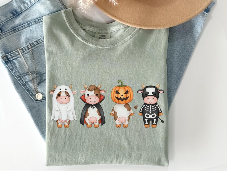 May include: Sage green t-shirt featuring four cartoon cows dressed in Halloween costumes: a ghost, a vampire, a jack-o'-lantern, and a skeleton. The shirt is laid on top of blue jeans and a tan hat.