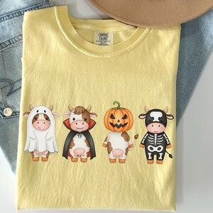 May include: Yellow Comfort Colors t-shirt featuring four cartoon cows in Halloween costumes: a ghost, a vampire, a jack-o'-lantern, and a skeleton. The shirt is laid flat on a light surface with a pair of blue jeans and a necklace.